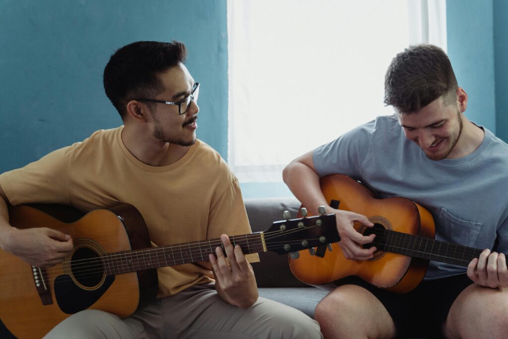 Two young men playing acoustic guitars together indoors, sharing a musical moment.