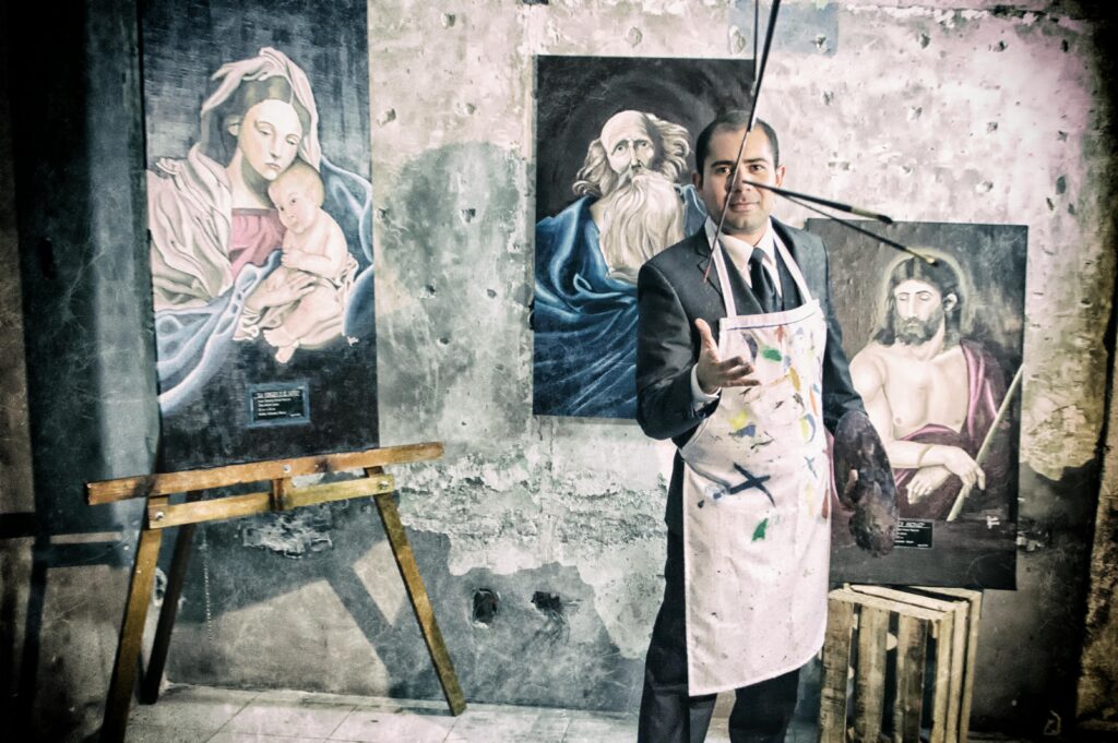 Artist in a suit and apron poses with Renaissance-style paintings in an old studio.