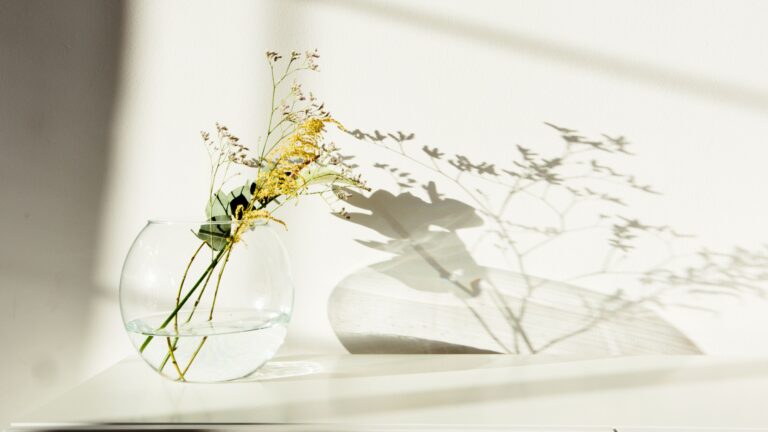 Simple and elegant glass vase with yellow flowers casting shadows on a white surface.